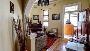 A spacious living room with a piano, a sofa, and a chair. There is a large window with natural light and a door leading outside.
