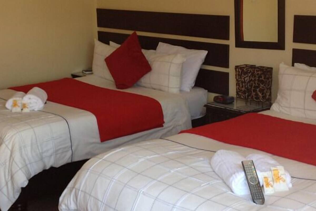 Habitación de hotel con dos camas, cada una con ropa blanca y detalles rojos, y una mesita de noche con una lámpara y un control remoto.