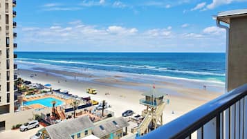 On peut admirer la plage depuis un balcon donnant sur une zone de villégiature avec une piscine, des parasols et un poste de surveillance pour les sauveteurs.