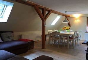 Une chambre à coucher confortable aménagée en mezzanine, avec un plafond en pente, des poutres en bois, un puits de lumière, un coin repas avec une table et des chaises, et un canapé agrémenté d’un plaid rouge.