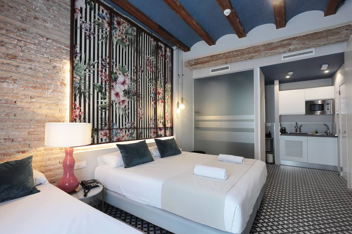 A hotel room with a bed, a red lamp, a floral wall panel, a kitchen area, and a patterned floor.