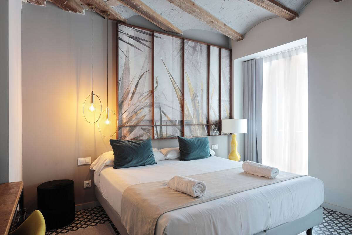 A modern hotel room with a large bed, a wooden ceiling, a painting on the wall, and a window with curtains.