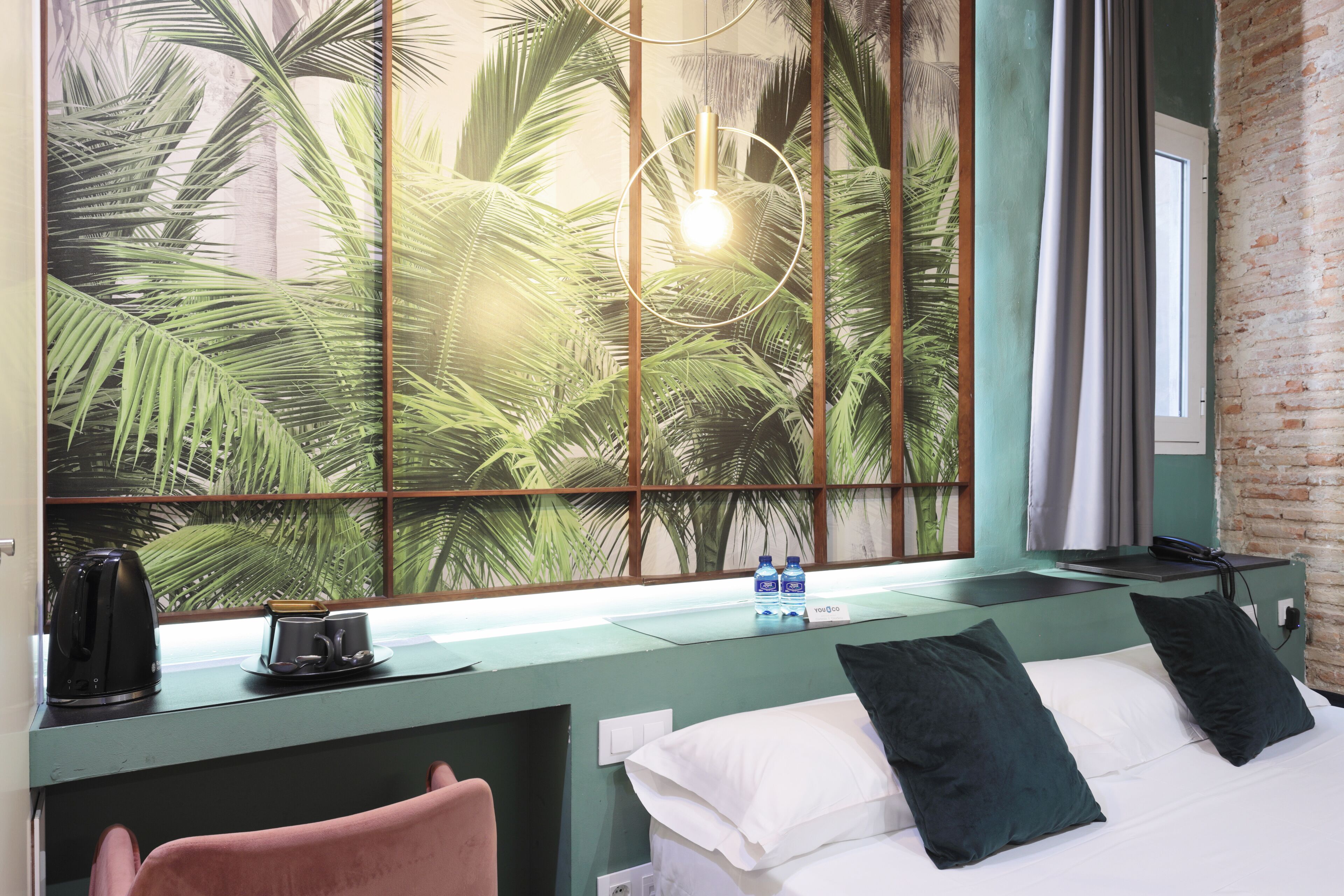 A hotel room with a bed, a desk with a lamp, a window with a view of palm trees, and a brick wall.