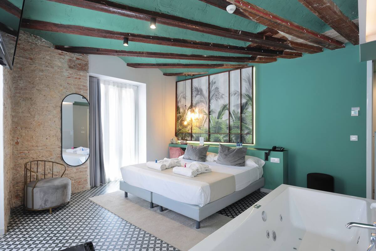 A bedroom with a bed, a large window, a bathtub, and a mirror.