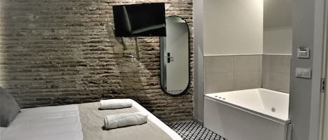 Habitación de hotel con pared de ladrillo, cama con ropa blanca, televisor de pantalla plana en la pared, un espejo grande y un baño separado con bañera y piso de baldosas.