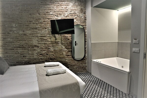 Habitación de hotel con pared de ladrillo, cama con ropa blanca, televisor de pantalla plana en la pared, un espejo grande y un baño separado con bañera y piso de baldosas.