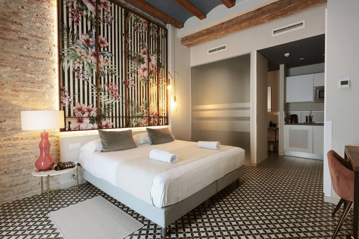 A modern hotel room with a large bed, a wooden headboard, a decorative wall panel, a small table with a lamp, and a kitchenette in the background.