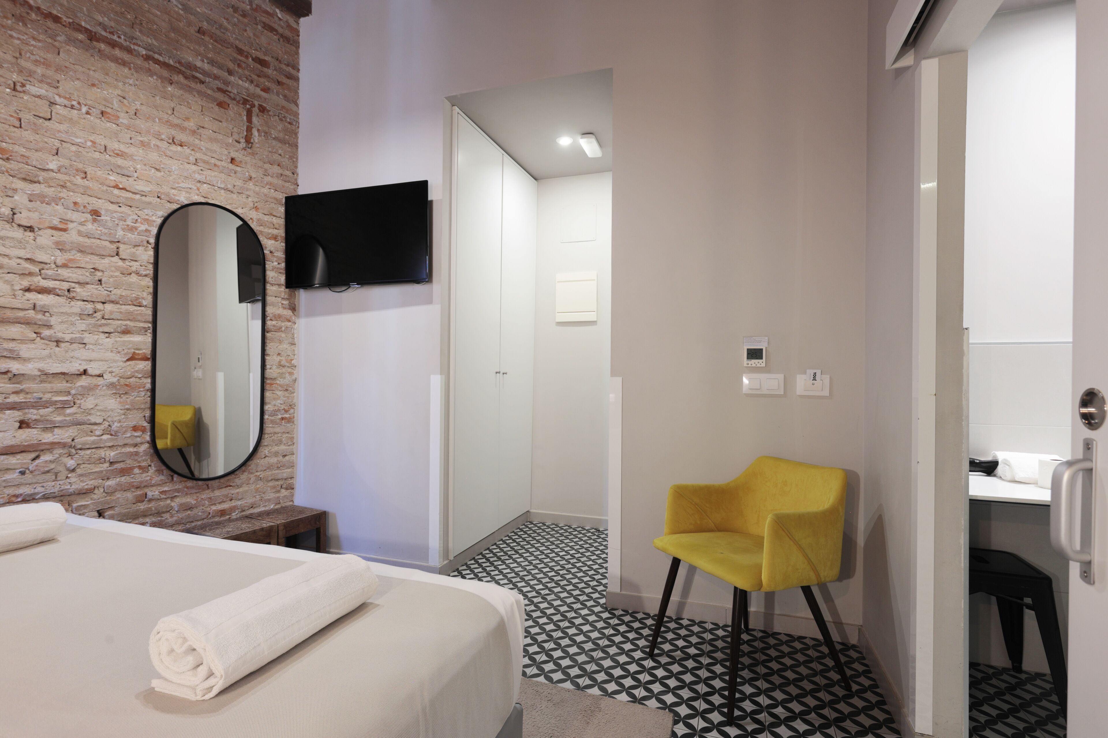 A hotel room with a brick wall, a bed with a rolled white towel, a yellow chair, a flat-screen TV, and a modern bathroom with a black and white patterned floor.