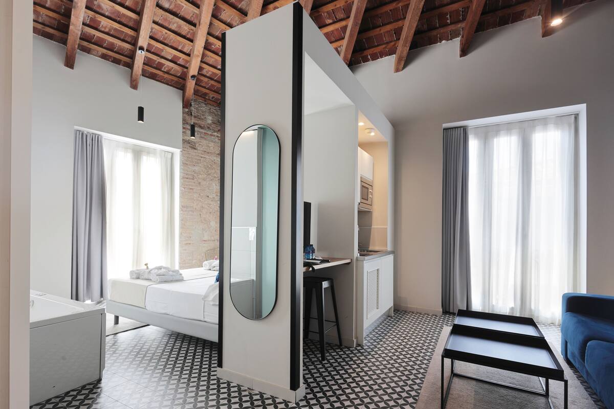 A modern hotel room with a bed, a desk, a chair, a large mirror, and a view of the city through the windows.