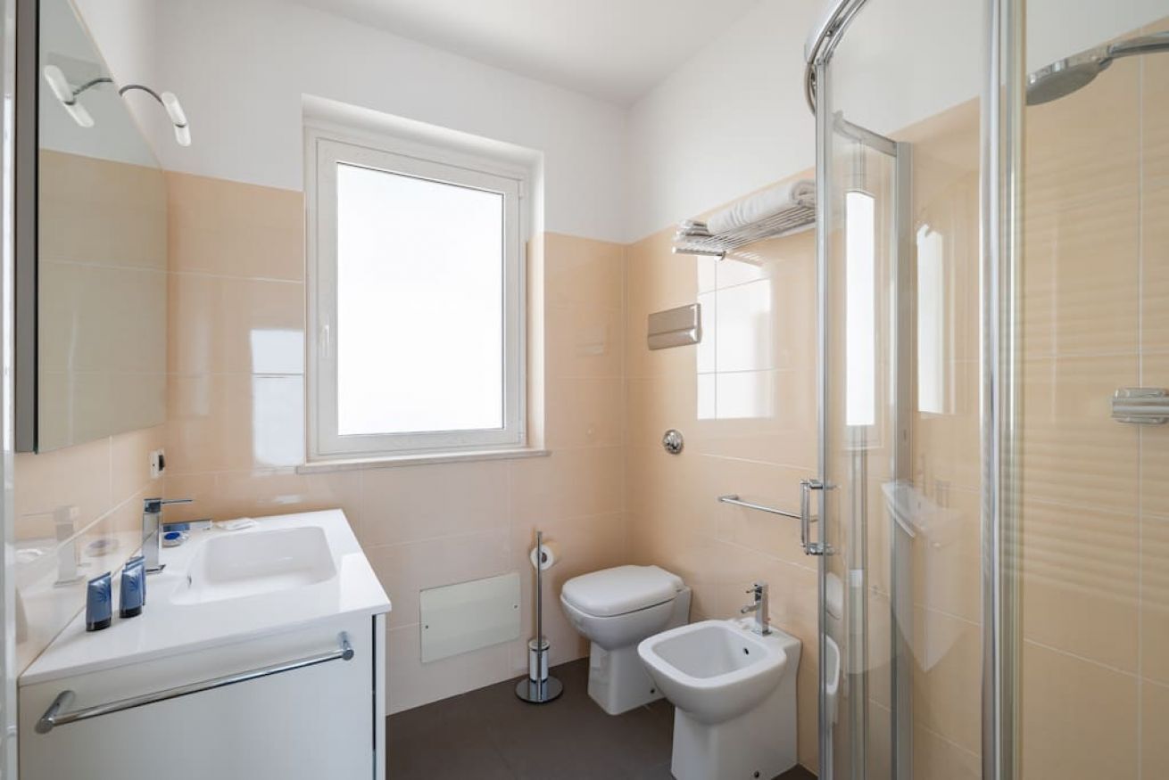 A modern bathroom with a large window, a sink, a toilet, a bidet, and a glass-enclosed shower area.