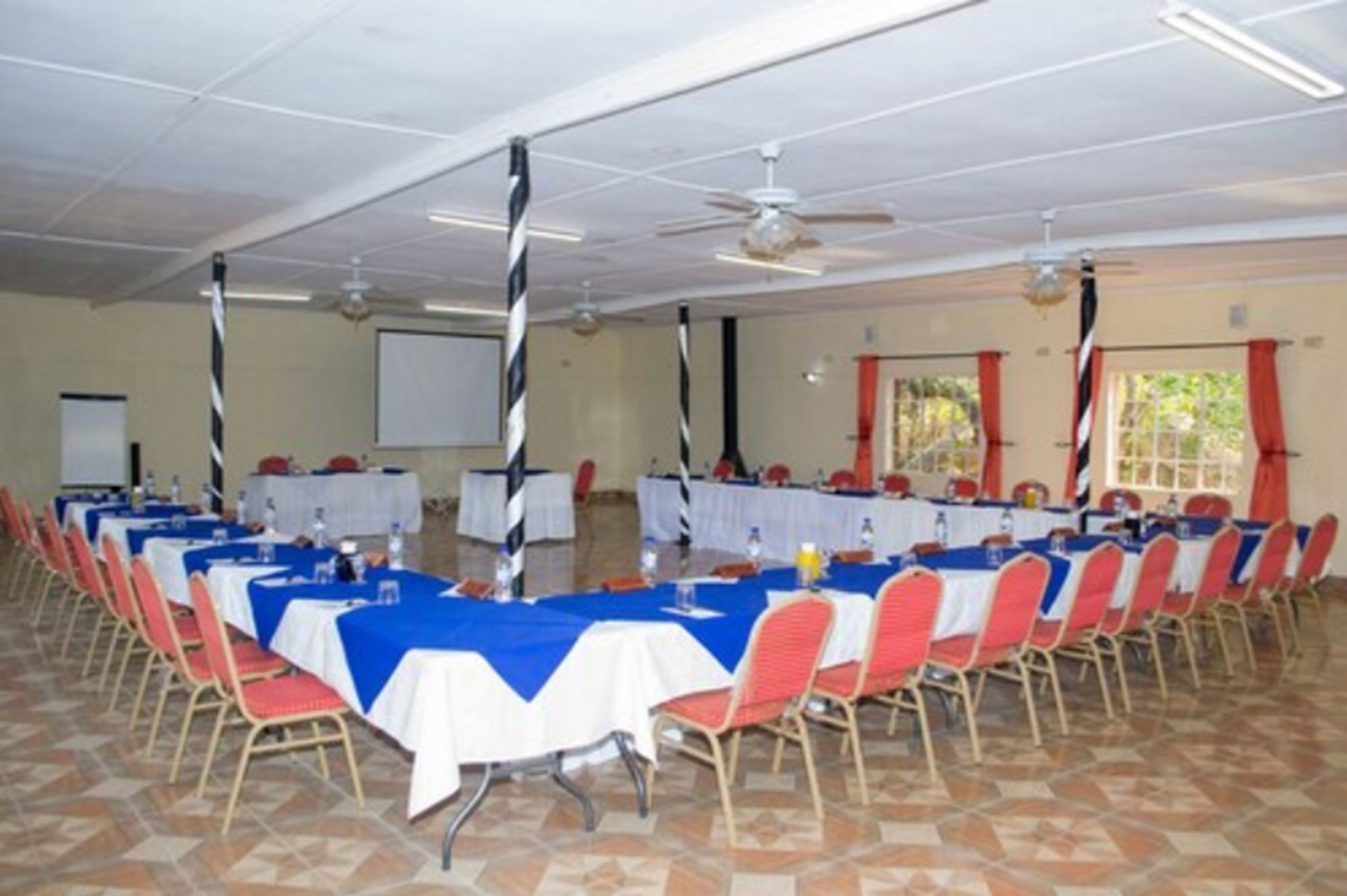 Un salón de conferencias con mesas dispuestas para una reunión, equipado con sillas, mesas y una pantalla de proyector.