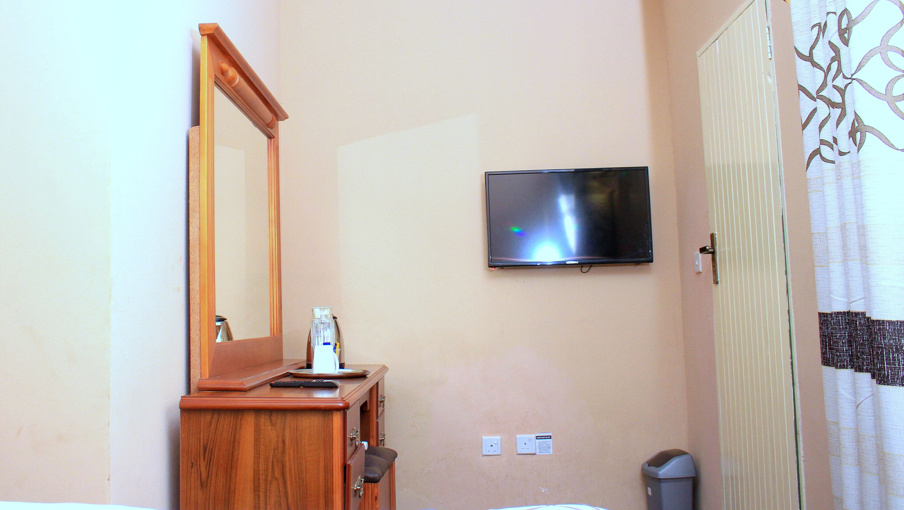 A room with a wooden dresser, a flat-screen TV mounted on the wall, a mirror, a door with a patterned curtain, and a trash bin.