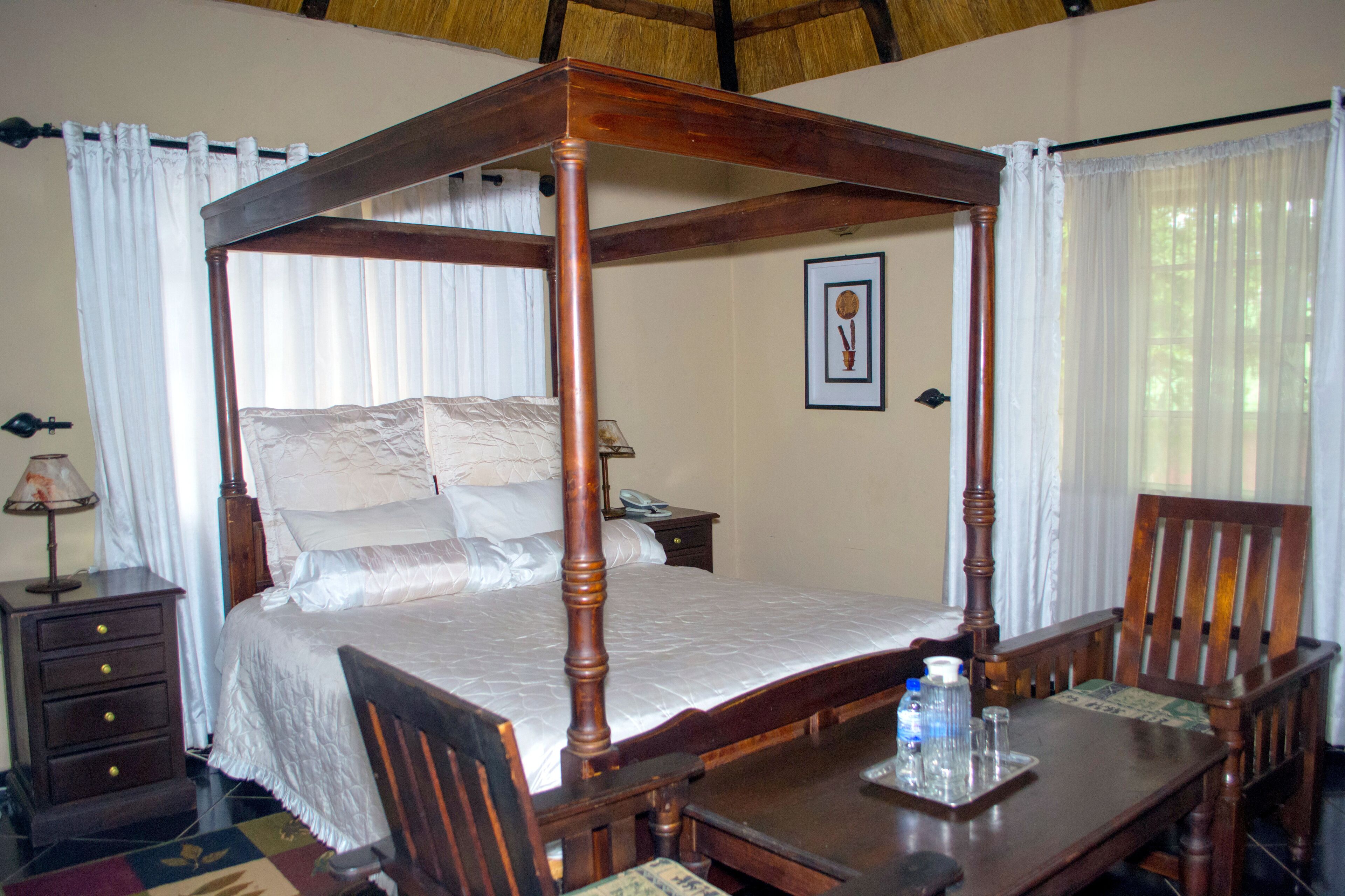 A four-poster bed with a wooden frame, a bedside table with a lamp, a chair, and a tray with a bottle and glasses.