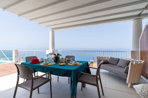 A covered outdoor dining area with a table set for two, chairs, and a view of the sea.