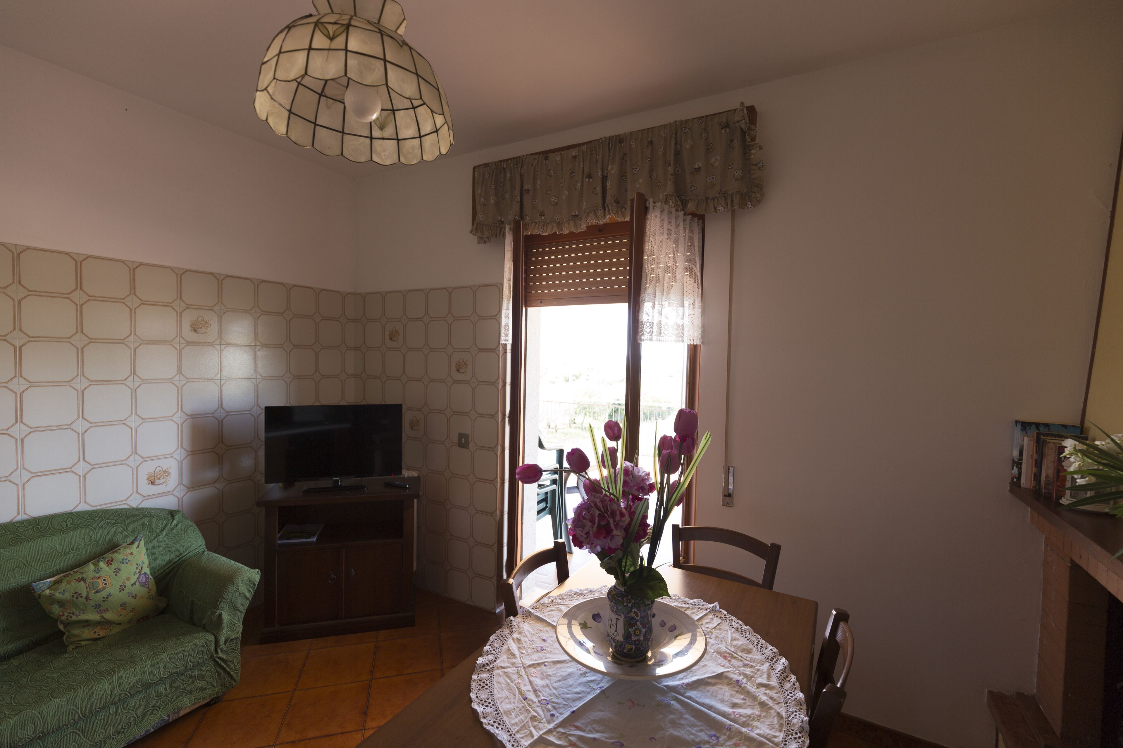 A living room with a green sofa, a TV on a stand, a dining table with a lace tablecloth, and a vase with flowers.
