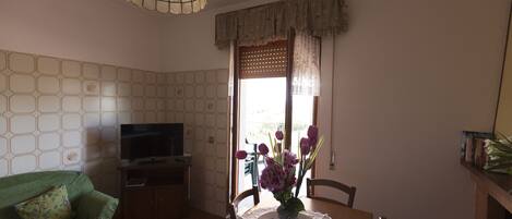 A living room with a green sofa, a TV on a stand, a dining table with a lace tablecloth, and a vase with flowers.