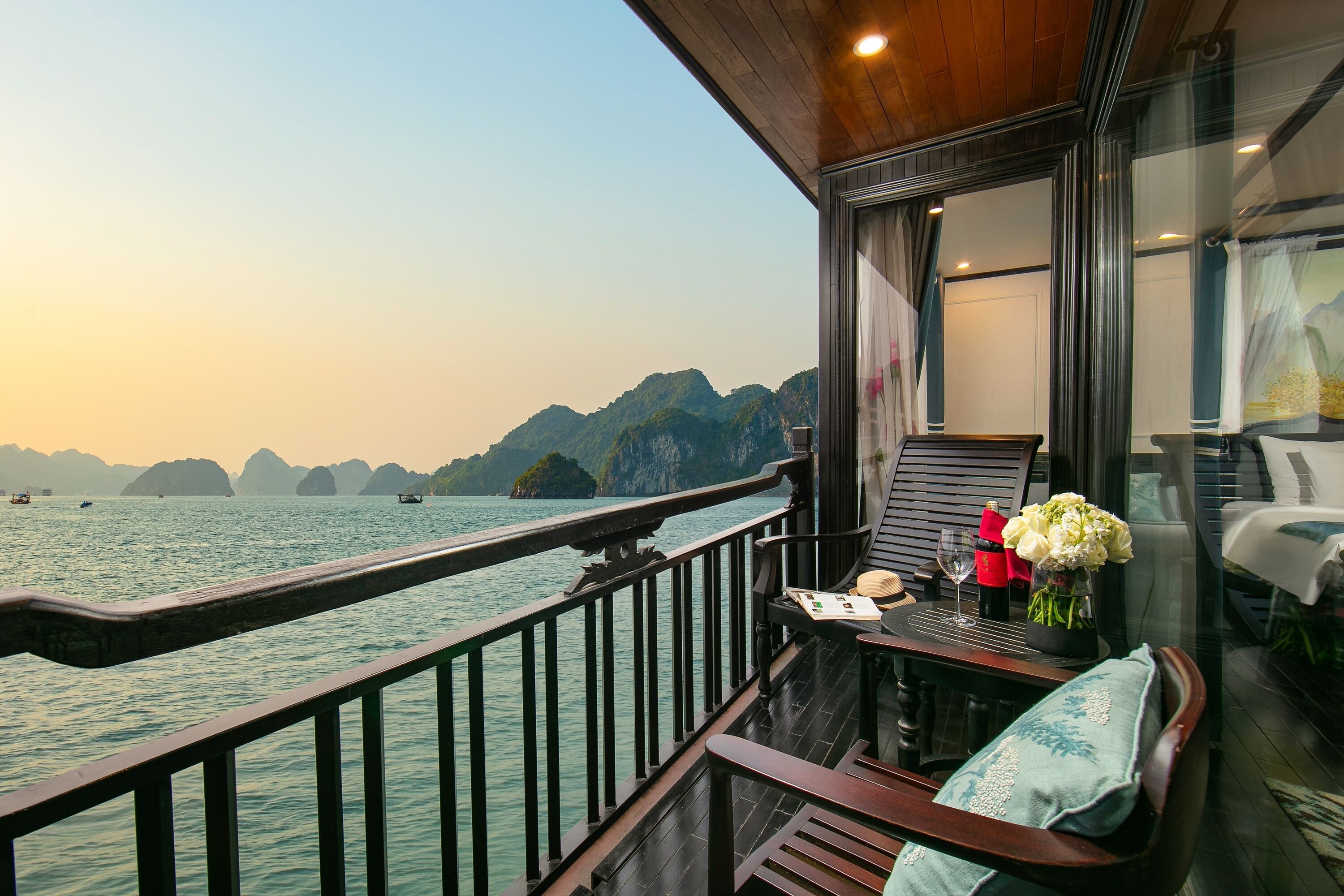 A balcony with a view of the water and mountains, wooden furniture, and a table with a vase of flowers.
