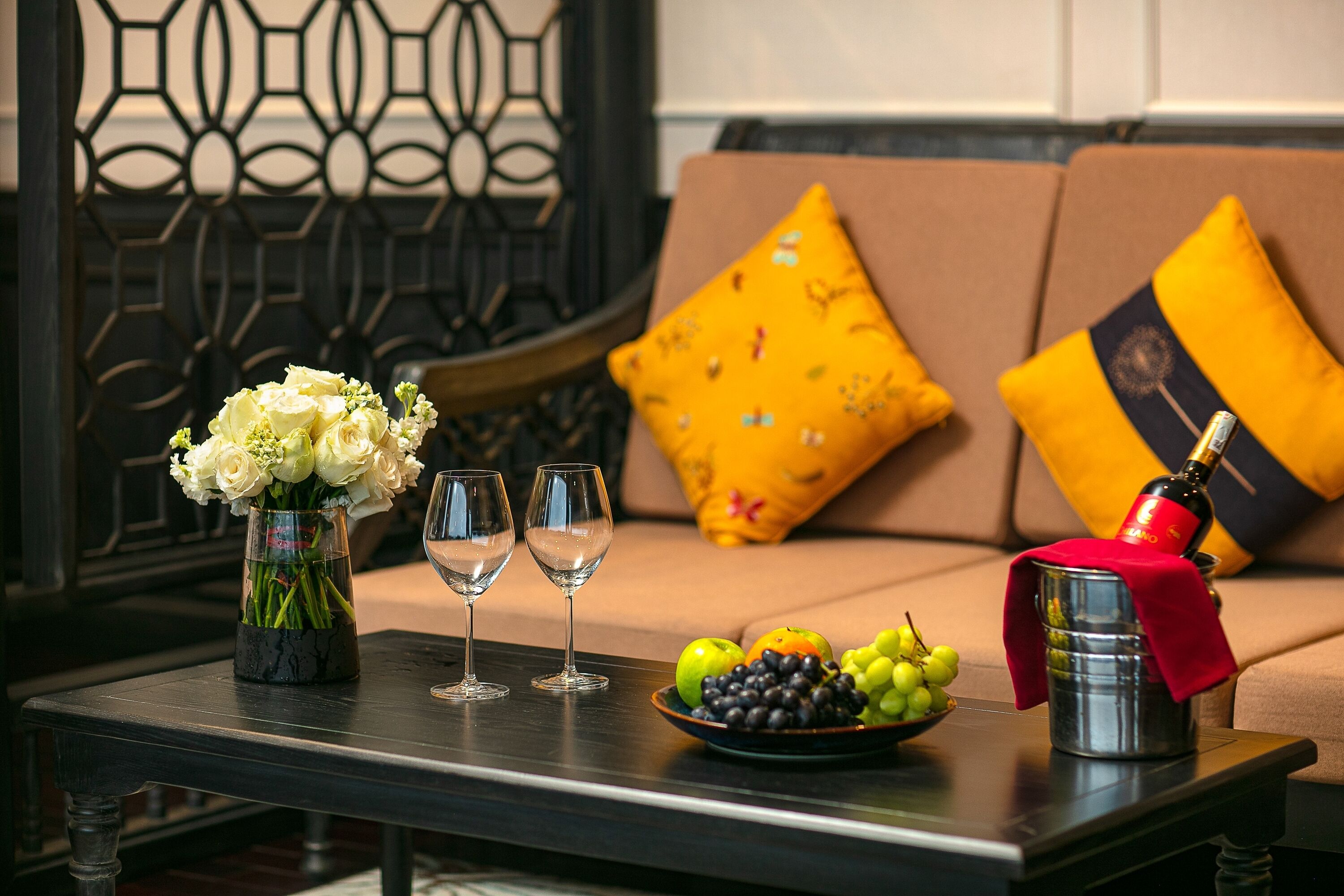Une table avec un vase de fleurs, deux verres à vin, un saladier de fruits et une bouteille de champagne.