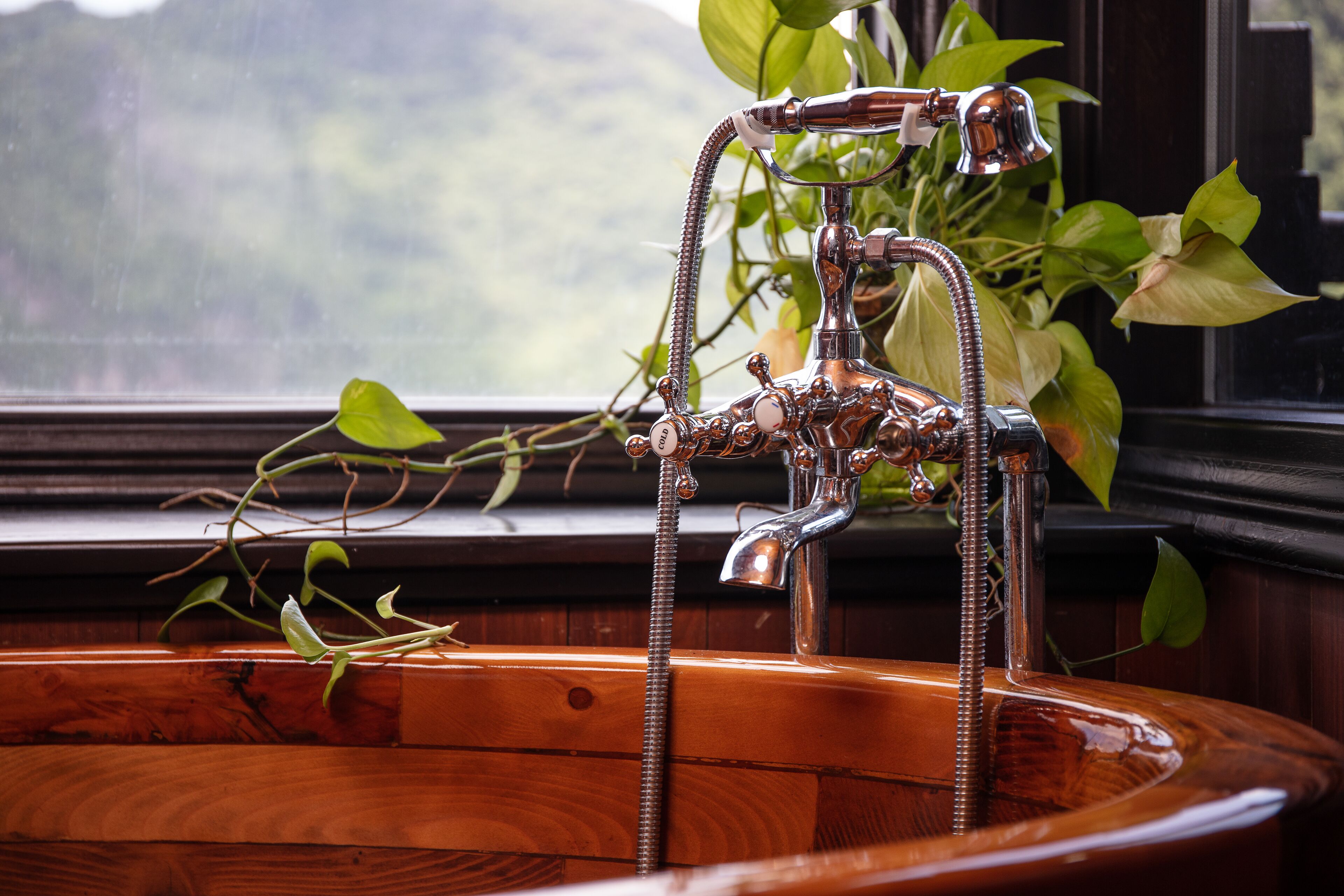 A wooden bathtub with a vintage faucet and a view of a forest through a window.