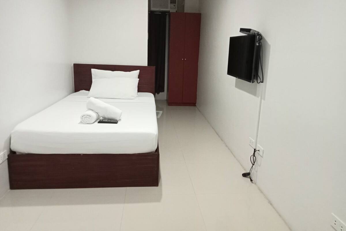 Una cama bien hecha con ropa blanca y almohadas, un cabecero de madera, un televisor montado en la pared y un armario rojo al fondo.
