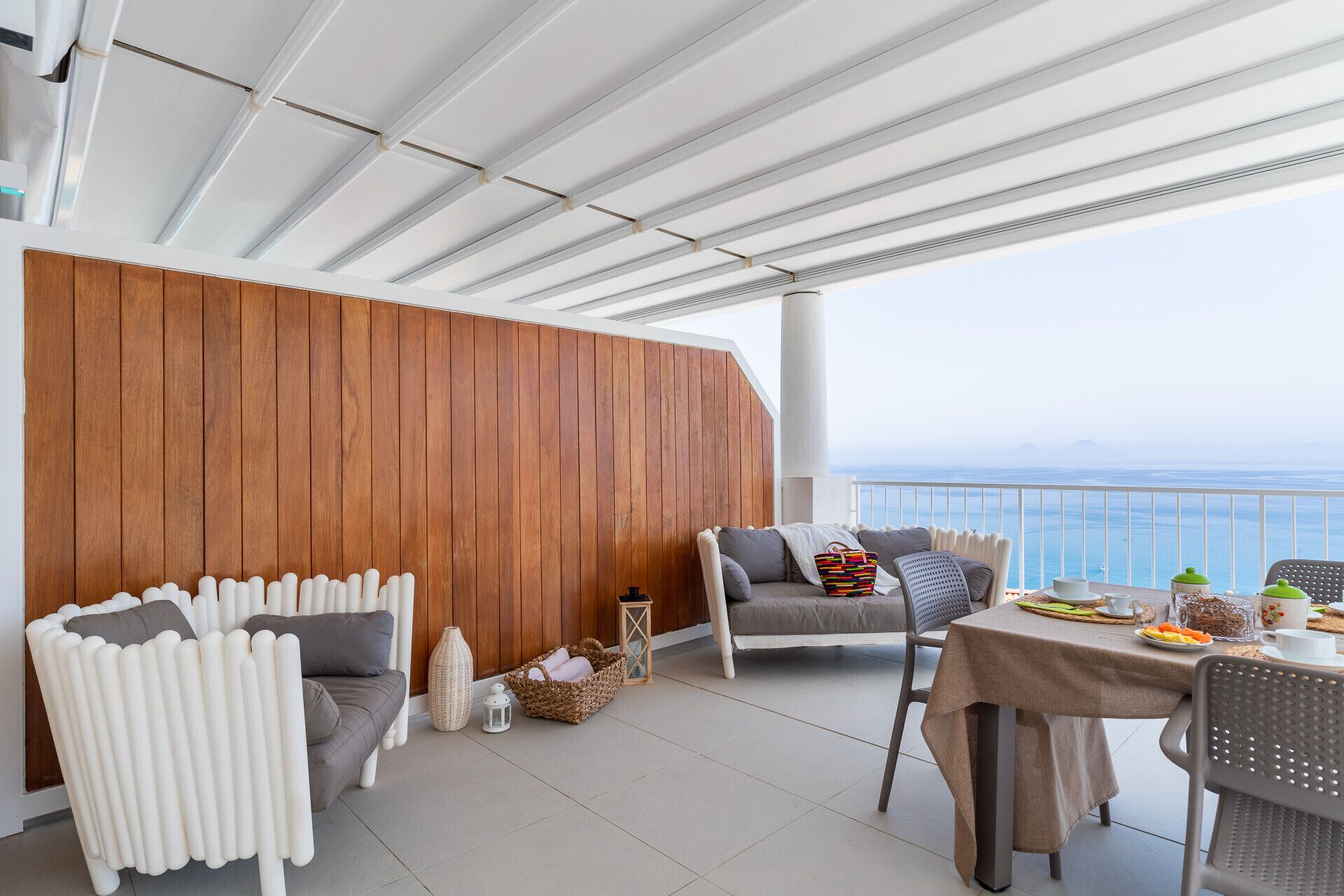 A balcony with a wooden panel wall, white chairs, a grey sofa, and a table set for dining.
