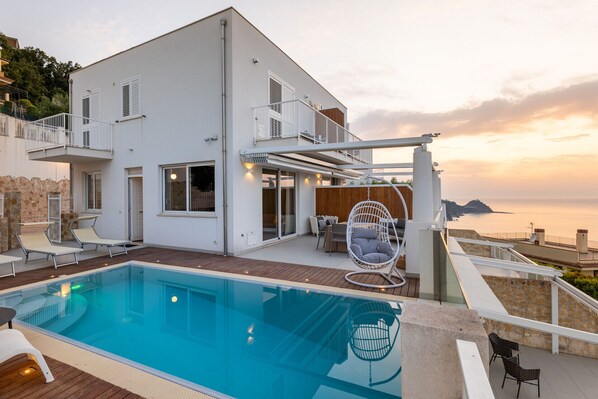 A modern two-story house with a swimming pool, outdoor seating, and a view of the sea.