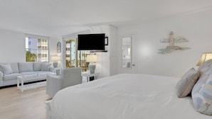 Une chambre d’hôtel avec un grand lit, un canapé, un fauteuil, une télévision et un balcon donnant sur des immeubles.