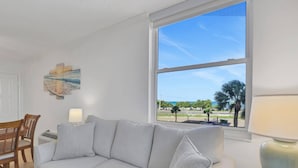 Un salon comprenant un canapé, une table à manger, des chaises, une lampe et une grande fenêtre donnant sur des palmiers et une plage.