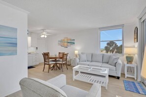 A living room with a white sofa, a coffee table, and a dining area with wooden chairs and a white table. - 3RD FLOOR STUDIO | POOL-VIEW LANAI | KING BED | FREE PARKING | STEPS TO BONITA BEACH (Bonita Springs)