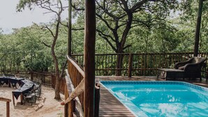 Una terraza de madera con piscina, rodeada de árboles y un área cercada con mobiliario exterior.