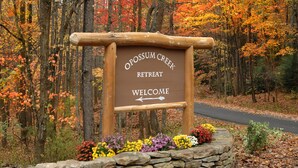 A wooden sign with "Opossum Creek Retreat Welcome" written on it, surrounded by colorful flowers and a stone wall.