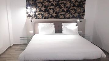 Une chambre avec un grand lit, une literie blanche, une tête de lit dotée de lampes murales et un papier peint orné de motifs de feuilles tropicales au-dessus du lit.