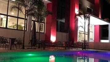 A nighttime pool area with a reflection of chairs and palm trees, surrounded by a building with red and white walls and large windows.