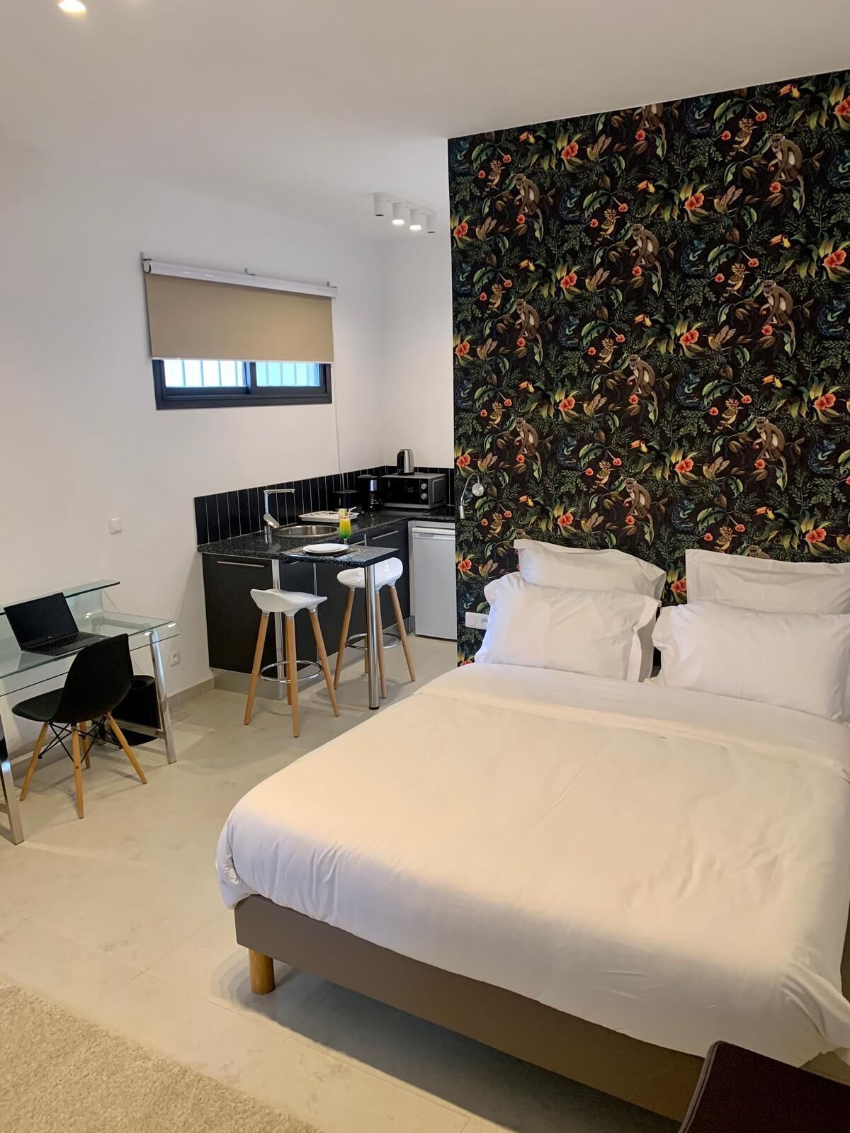 A bedroom with a bed, a desk with a laptop, a chair, and a kitchenette in the background.