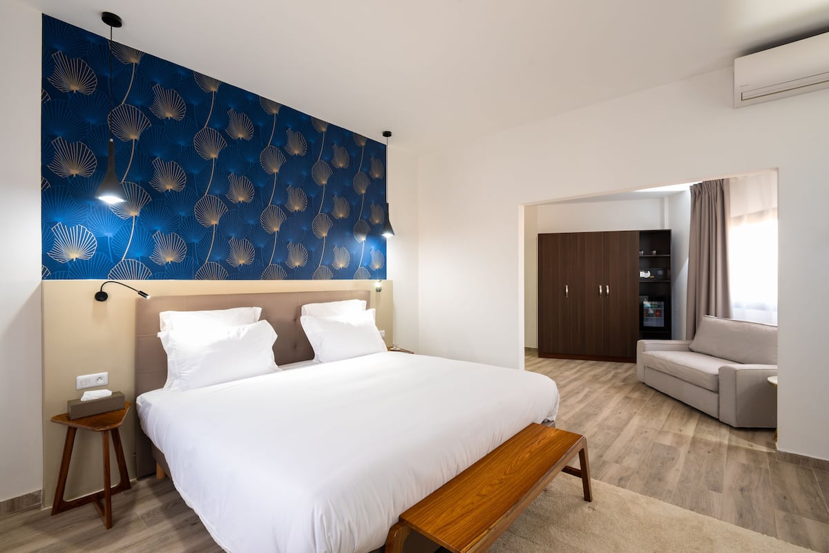 Una habitación de hotel moderna con una cama grande, un banco de madera, una mesita de noche, un armario y una zona de estar con sofá.