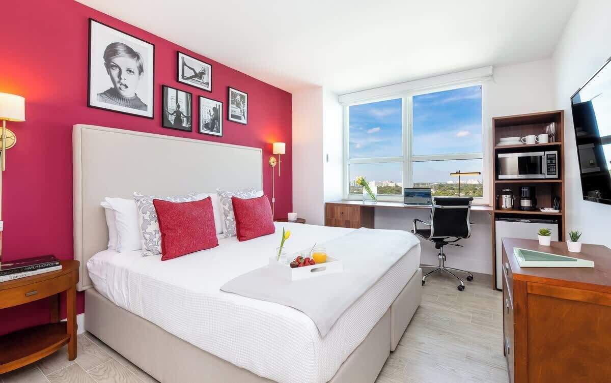A hotel room with a large bed, a desk, a chair, a TV, and a window with a city view.