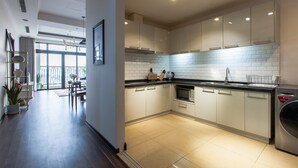 Una cuina moderna amb armaris blancs, un taulell negre i un vàter d'acer inoxidable. Es veu una zona de dinar amb una taula i caires a través d'una porta oberta.