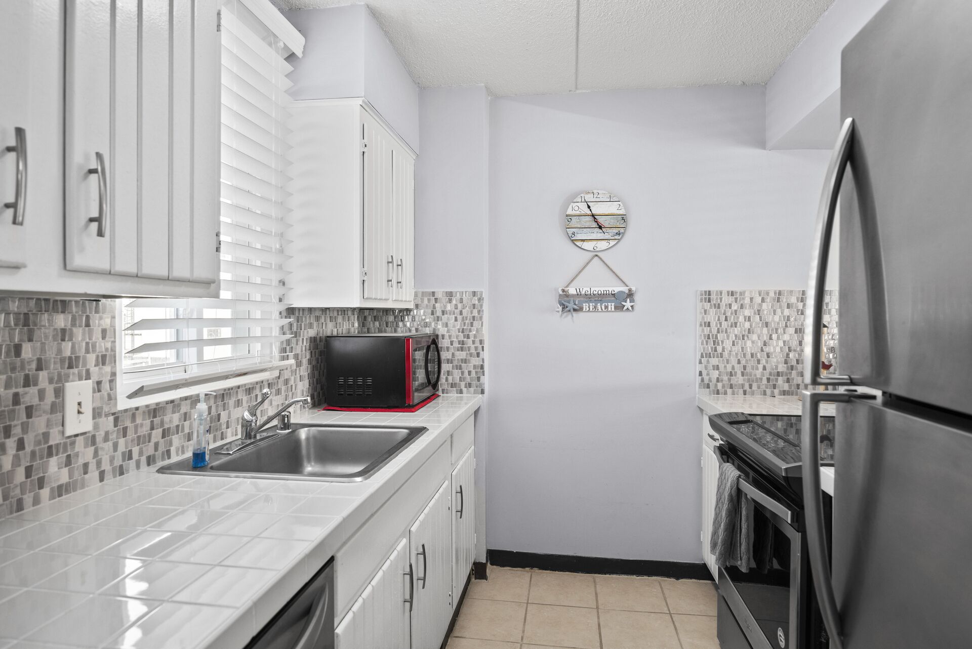 A compact kitchen with white cabinets, a black coffee maker, a stainless steel refrigerator, and a wall clock.