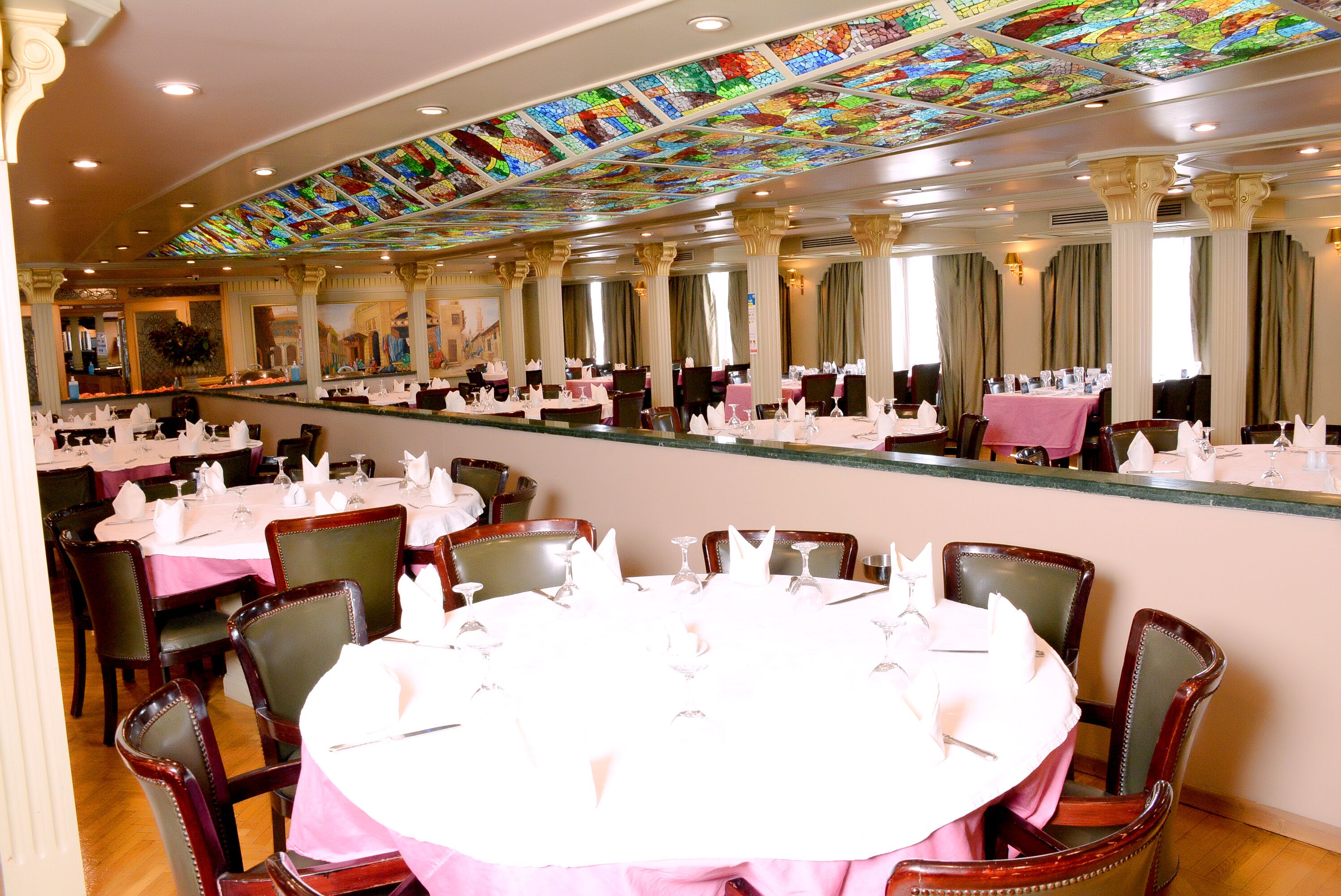 Ein Bankettsaal mit runden Tischen, Stühlen und einer Decke aus Buntglas.