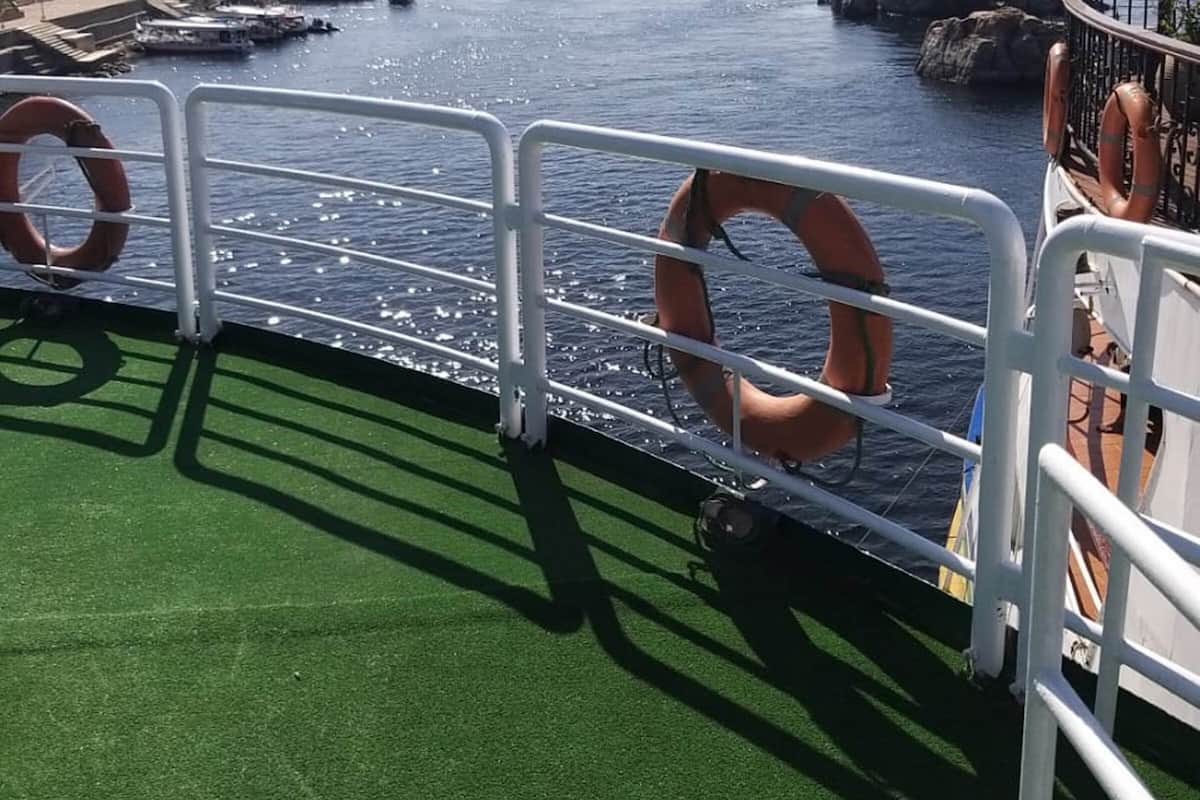Een dek met een reling, drijvers en uitzicht op een haven met boten en gebouwen.