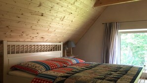 Ein Schlafzimmer mit schrägen Wänden, einem Bett mit gemusterten Betttextilien, einem Nachttisch mit Lampe und einem Fenster mit Vorhängen.