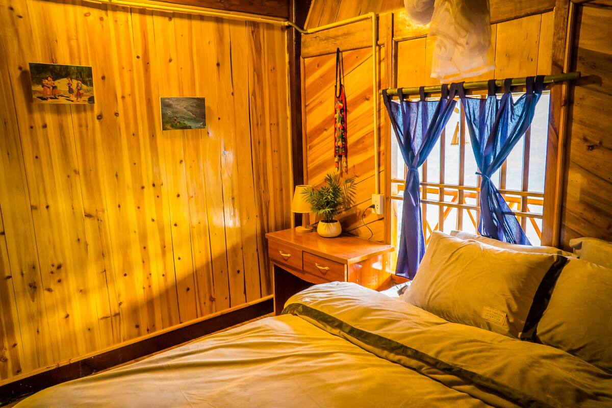 A wooden room with a bed, a nightstand, a lamp, and a window with curtains.