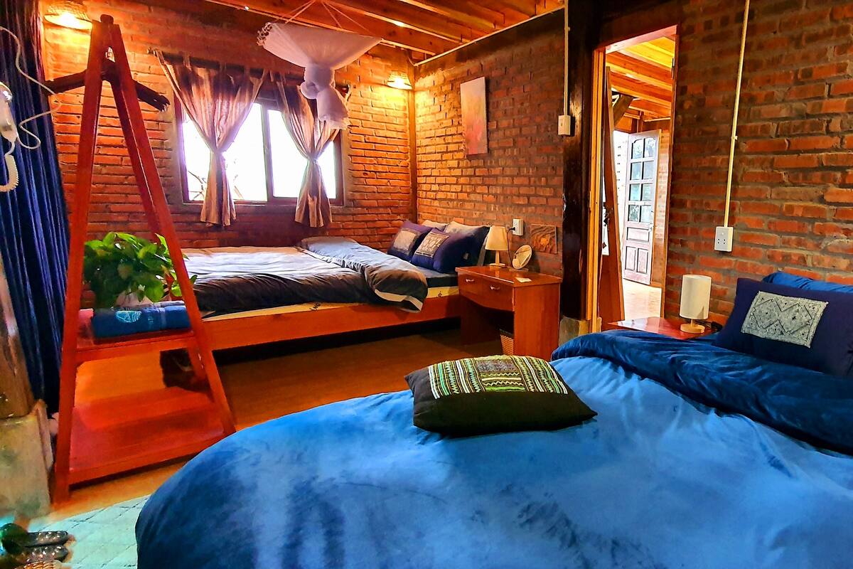 A rustic room with a brick wall, a wooden ladder, a bed with blue bedding, and a small table with a lamp.