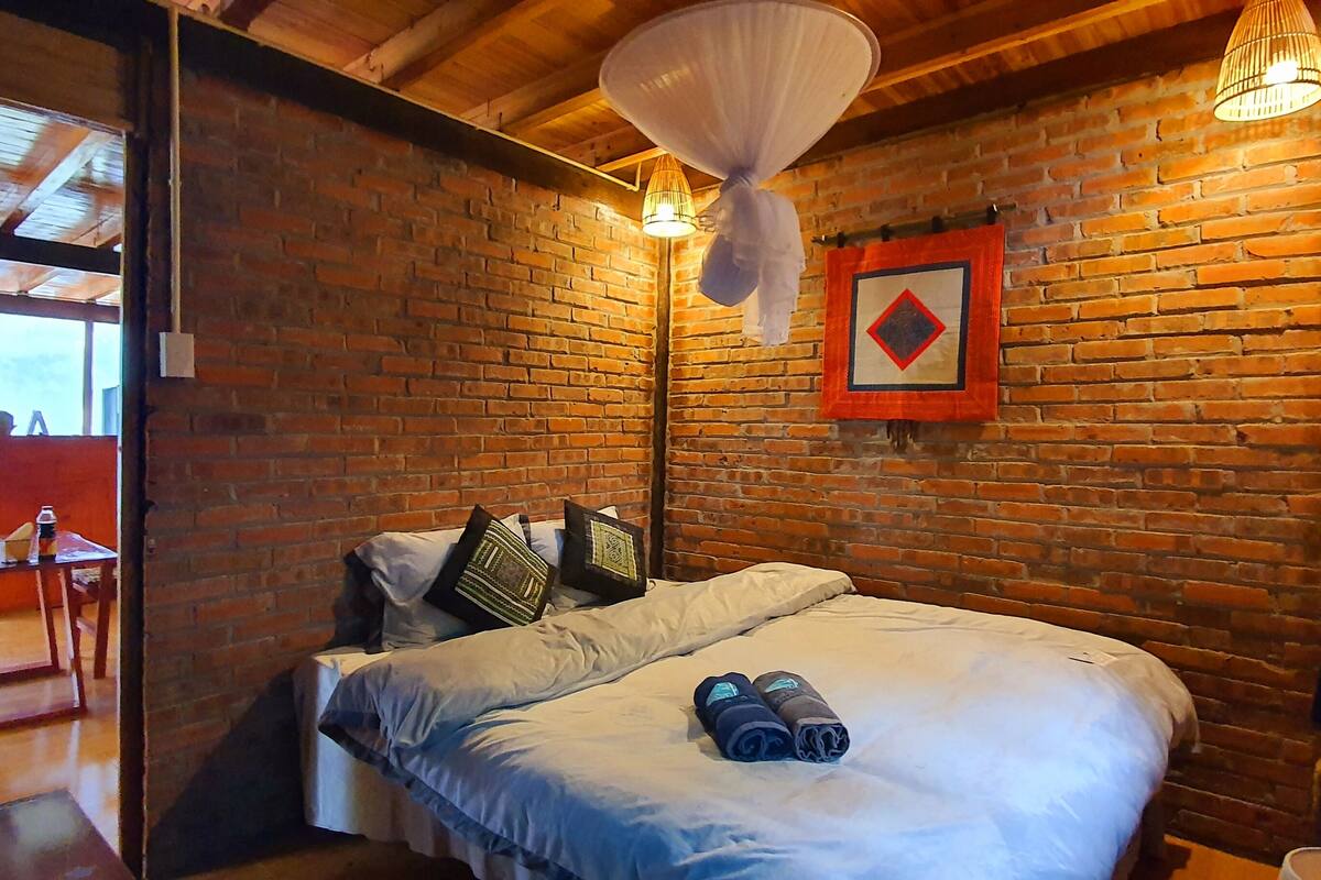 A cozy room with a brick wall, a bed with pillows, a hanging light fixture, and a window with a view.