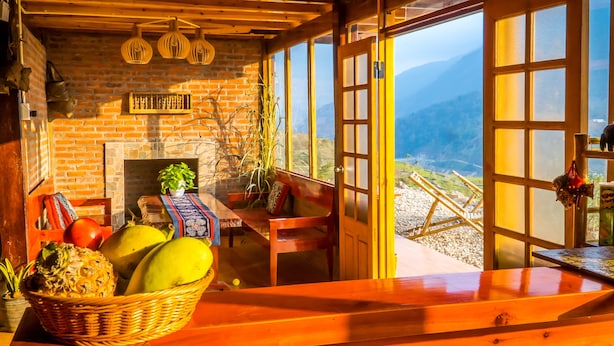 A rustic wooden interior with a brick fireplace, wooden ceiling beams, and a view of mountains through large windows. - The Little H'mong House (Ta Van)