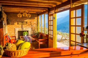 A rustic wooden interior with a brick fireplace, wooden ceiling beams, and a view of mountains through large windows. - The Little H'mong House (Ta Van)
