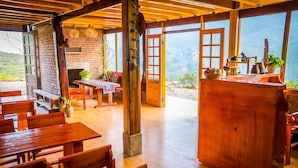 A rustic wooden interior with a dining area, open doors leading to a scenic view, and a brick fireplace. - The Little H'mong House (Ta Van)