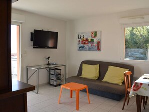 A living room with a sofa, a television, a desk with a computer, and a window with a view of greenery.