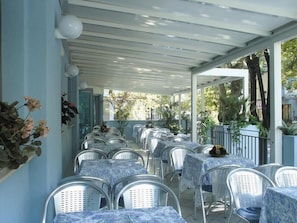 A covered outdoor dining area with rows of tables and chairs, surrounded by potted plants and trees. - Hotel Feldberg (Rimini)