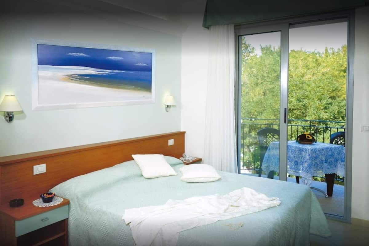 A bedroom with a large bed, two bedside lamps, a painting of a beach, and a balcony with a table and chairs.